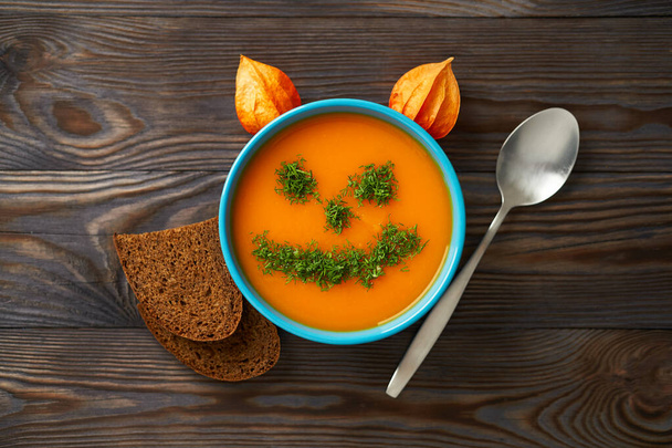 Autumn cream soup garnished with dill in the form of funny faces Halloween pumpkins. Healthy pumpkin and carrot soup puree in a blue bowl on a wooden background. Halloween holiday concept, top view. - Photo, Image