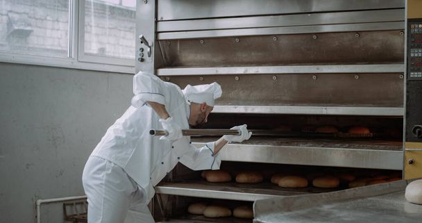 クローズアップパン屋はオーブンからベーキングパンを取り出し、特別なトーチを使用して棚にアンロード - 写真・画像