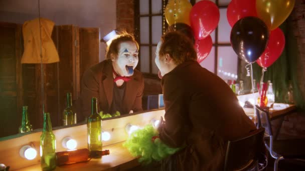 Een gekke man clown lacht zichzelf uit in de spiegel met zijn blauwe geschilderde tong - verknoeit zijn haar en wordt gek - Video