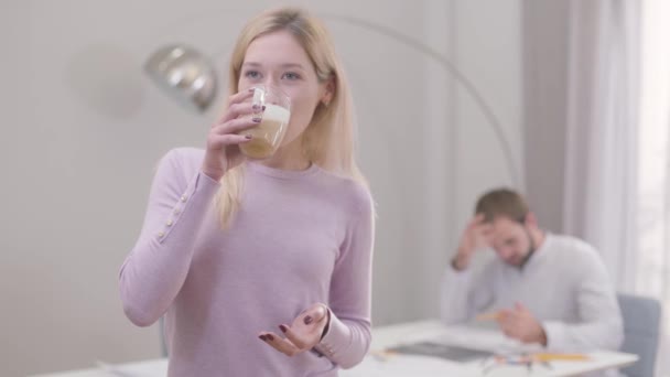 Portrait of young blond woman drinking cappuccino or latte and smiling. Professional architects having break. Her male colleague drawing blueprints at the background. - Footage, Video