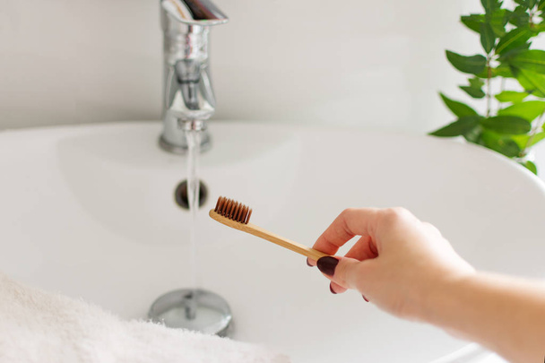 Mano de mujer usando un cepillo de dientes de bambú bio-degradable en un baño interior blanco. Decoración de la planta verde en el fondo
. - Foto, imagen