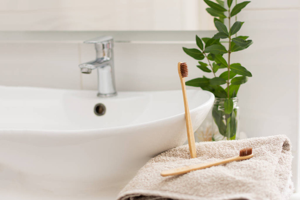 Dos cepillos de dientes de bambú bio-degradables y compostables sobre una toalla en el interior de un baño blanco. Decoración de la planta verde en el fondo
. - Foto, imagen