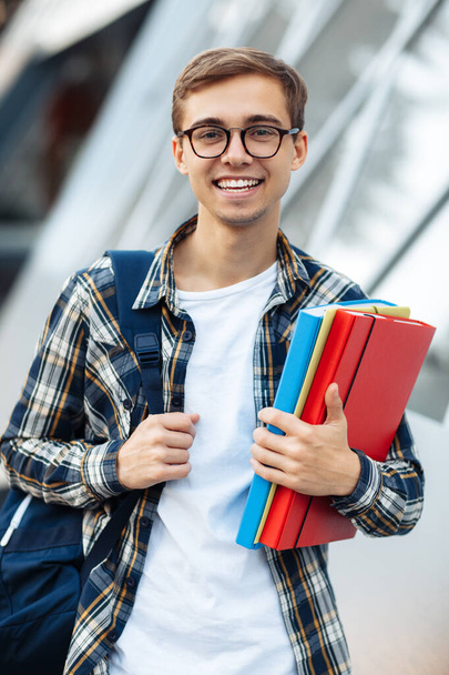 Cheerful young male student in casual outfit with bright smile holding folders while standing on the street. - 写真・画像