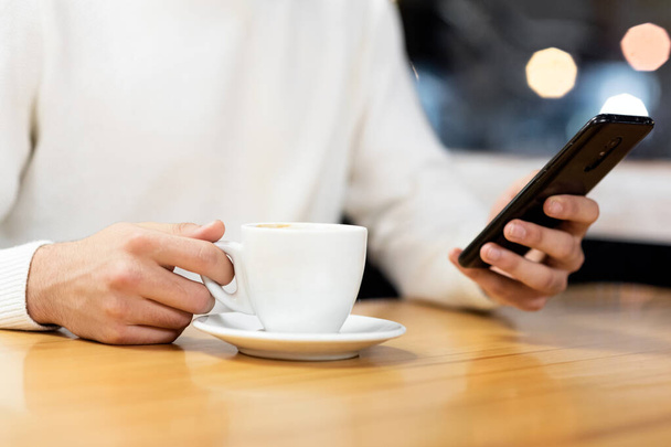 Молодий чоловік, що сидить у кафе, користується мобільним телефоном і випиває каву. Кавказький самець з чашкою кави і читанням текстового повідомлення на своєму смартфоні. - Фото, зображення