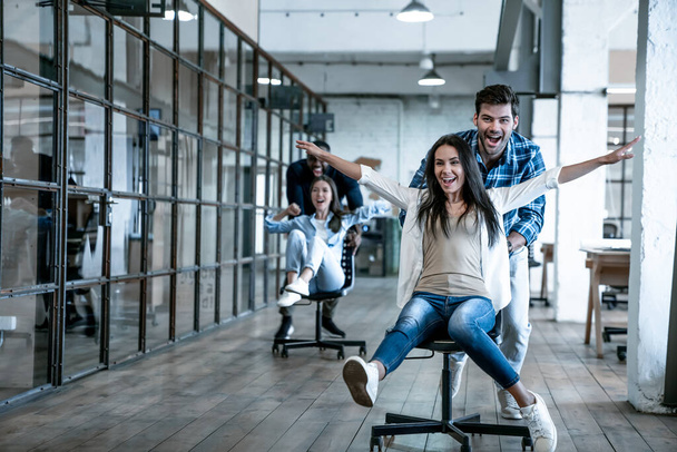 Δούλεψε σκληρά. Τέσσερα νεαρά χαρούμενα άτομα των επιχειρήσεων σε smart casual φορούν διασκεδάζοντας ενώ αγωνίζονται σε καρέκλες γραφείου και χαμογελώντας - Φωτογραφία, εικόνα