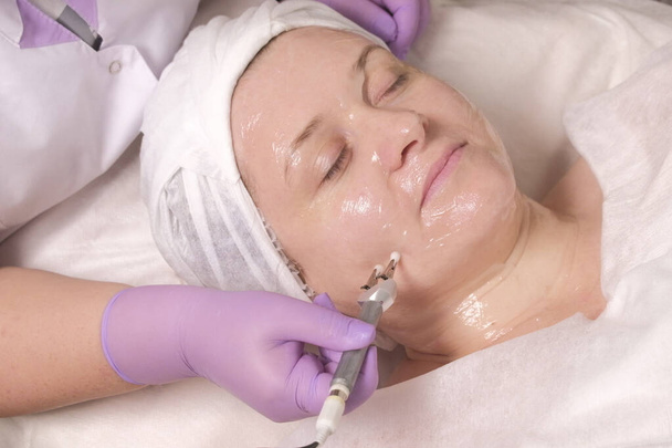 Hardver kozmetológia. Öregedésgátló eljárás a nők bőrére. Átlátszó zselé egy nő arcán. Az elektróda egy lila kesztyűs kozmetikus kezében. Lány egy szépségszalonban. - Fotó, kép