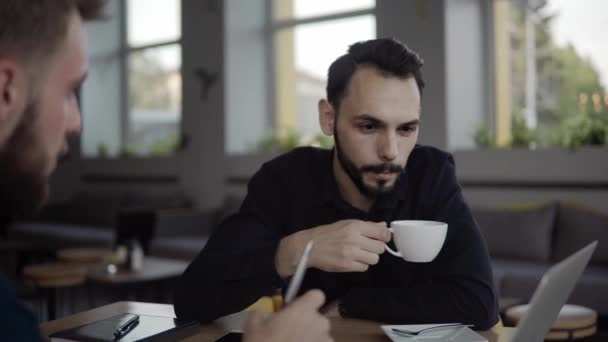 Two cheerful business mans discussing something and smiling while sitting at the cafe with a laptop and phones. Two young successful colleagues in discussing organization moments by table in cafe. - Materiał filmowy, wideo
