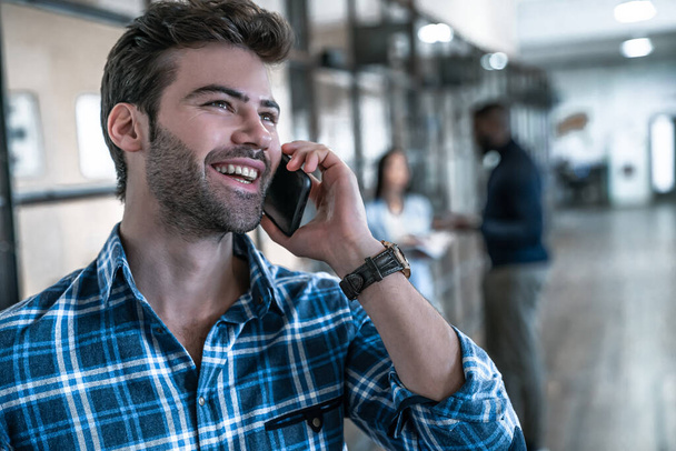 Conversa de negócios rápida. Bonito jovem sorrindo homem falando ao telefone e gesticulando enquanto seus colegas de pé no fundo
 - Foto, Imagem