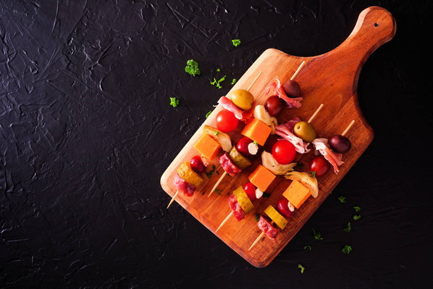 Skewered party appetizers with meat, cheeses and pickles on a serving board. Top view on a dark stone background. - Foto, Bild