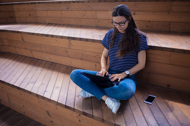 A serious brunette student girl , sits on wooden steps, with an open laptop, typing on a keyboard, learns online. - Photo, Image
