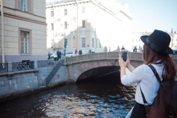 Tourist in the city takes a photo on smartphone. Young woman in black hat and white shirt. - Photo, image