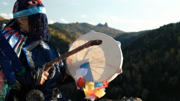 An old shaman in a bright national costume enters a trance banging a tambourine on the top of a mountain. - Footage, Video