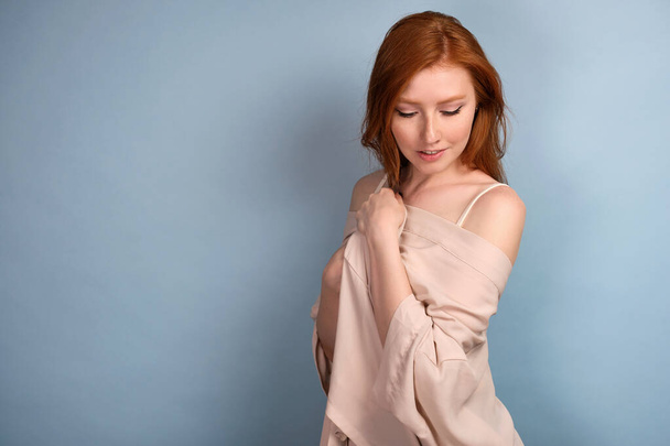 Red-haired girl on a blue background stands holding a dressing gown lowered from her shoulders, looks down - Foto, imagen