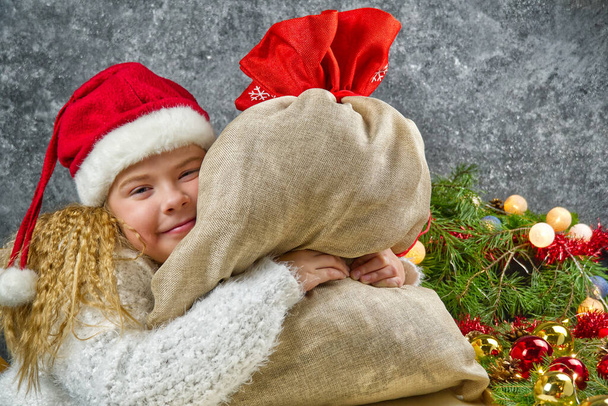 Niña rubia en un sombrero rojo gnomos con bolsa de santas en anticipación de un milagro de Navidad abraza una bolsa con regalos de Navidad
 - Foto, imagen