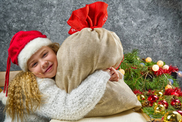 Маленька блондинка в гномах червоний капелюх з сумкою Сантас в очікуванні різдвяного дива обіймає сумку з різдвяними подарунками
 - Фото, зображення
