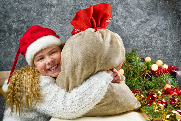 Malá blondýnka dívka v gnómy červený klobouk s sáčkem santas v očekávání vánočního zázraku objetí taška s vánočními dárky - Fotografie, Obrázek