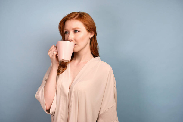 A red-haired girl in a light robe stands half a turn on a blue background and takes a sip from a light mug. - Foto, Imagen