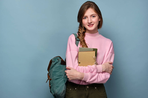 A blue-eyed girl in a pink sweater stands on a blue background with a backpack and books in her hands. - Fotoğraf, Görsel