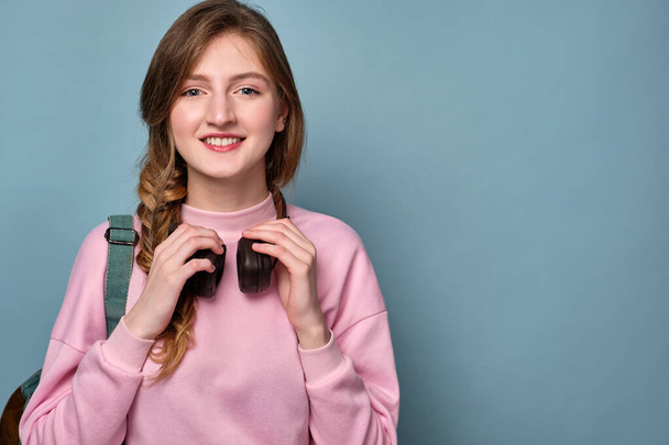 A girl with blue eyes and a braid in a pink sweater holds headphones on her neck and smiles. - Foto, afbeelding