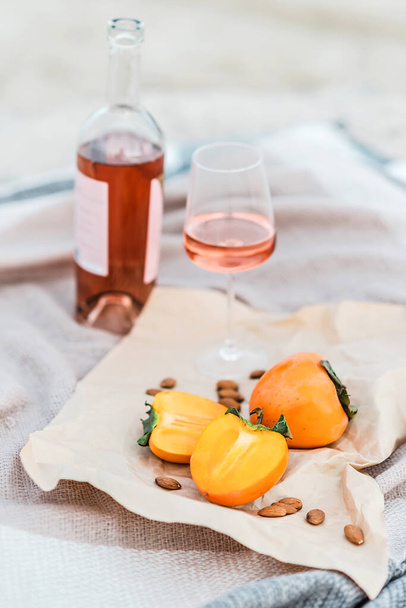 Picnic on the beach with persimmons, almond and bottle of rose wine on beige blanket. - Photo, Image