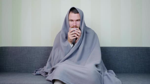 Nemocný muž přikrytý přikrývkou pije horký šálek čaje. Rozrušený mladík sedí na gauči s vysokou teplotou. Akutní respirační virová infekce. Nemoc, chřipka. Samoléčba doma. 4k - Záběry, video