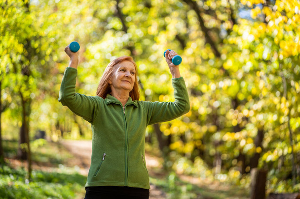 Mujer mayor está haciendo ejercicio con pesas en el parque en el día soleado
. - Foto, Imagen