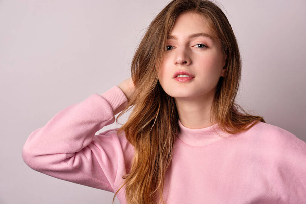 A blonde girl in a pink sweatshirt stands on a white background and puts her hand behind her head looking at the camera - Foto, afbeelding
