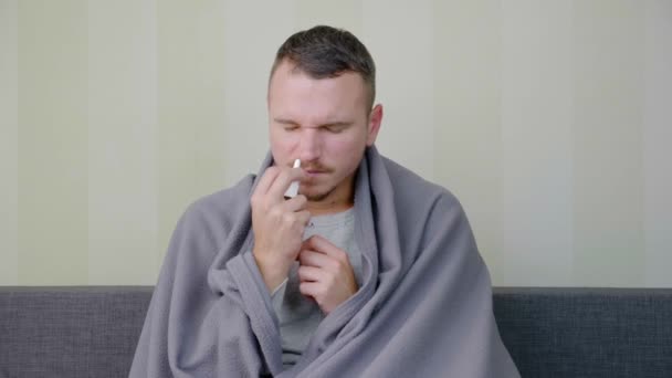 Kranker Mann mit Decke wird mit Nasenspray behandelt. Aufgebrachter junger Kerl sitzt auf der Couch bei hoher Temperatur. akute Infektion der Atemwege. Erkältungsgrippe. Selbstmedikation zu Hause 4k - Filmmaterial, Video