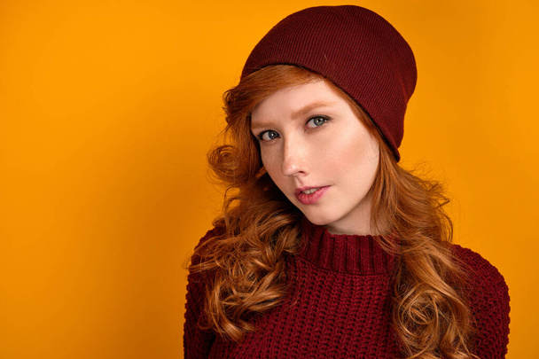 Close portrait of curly red-haired girl in a burgundy sweater and hat, standing on an orange background. - Foto, imagen