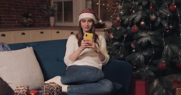 Beautiful Smiling Caucasian Blond Girl Wearing Santas Hat Sitting on a Sofa at Christmas Home Background Holding a Smartphone Texting Sending and Reading Messages. Concept of Holidays and New Year. - Filmati, video