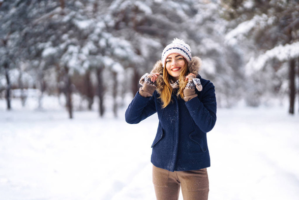 Portrait of young beautiful girl with long hair, in a blue jacket and knitted hat and mittens posing in sunny winter day. Fashion young woman in the winter forest. Christmas, winter holidays concept.  - Fotó, kép