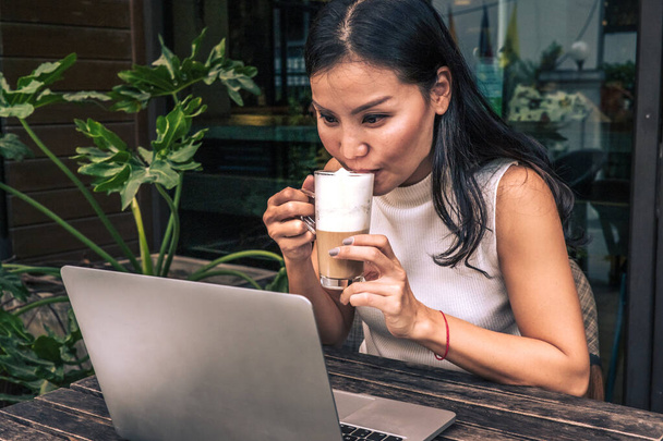 Mulher asiática bebendo um café enquanto assiste um laptop
 - Foto, Imagem