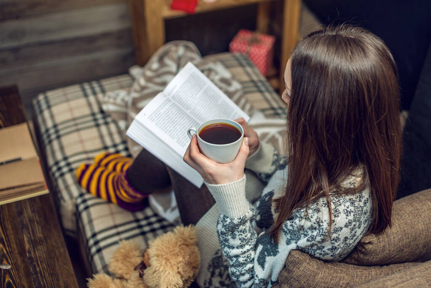 Femme en pull lisant un livre avec une tasse de café le soir dans une ambiance chaleureuse de Noël. Confortable nouvelle humeur de l'année
 - Photo, image