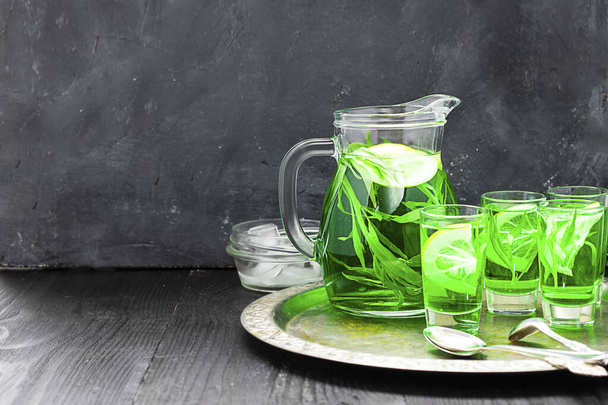 Summer cold green drink with tarragon, mint and lemon in glass decanter and small shots with ice, one in focus in focus. Sparkling beverage, alcohol drink on dark background. Horizontal, copy space - 写真・画像