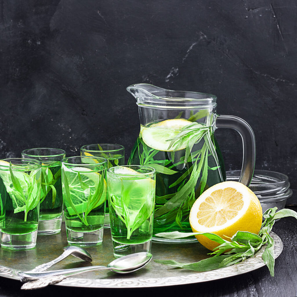 Summer cold green drink with tarragon, mint and lemon in glass decanter and small shots with ice, one in focus in focus. Sparkling beverage, alcohol drink on dark background. Square with copy space - Zdjęcie, obraz