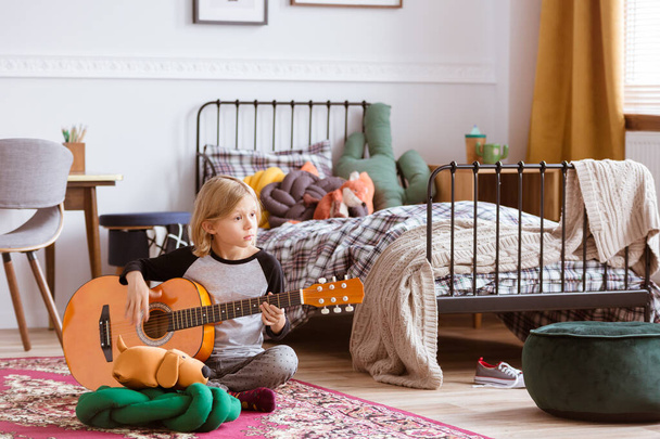 Cute blond girl sitting on the floor of her trendy bedroom,learning how to play a guitar - 写真・画像