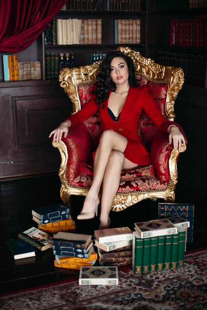 Portret van brunette in rode jurk zittend op fauteuil in bibliotheek interieur. - Foto, afbeelding