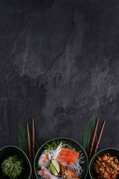 Sushi auf dunklem Schieferhintergrund. Wakame, Salat mit Meeresfrüchten, Lachs, Jakobsmuscheln, Aal und Garnelen Sashimi serviert mit Daikon Rettich. vertikal, Kopierraum für Text. Restaurantkarte. traditionelle japanische Küche - Foto, Bild