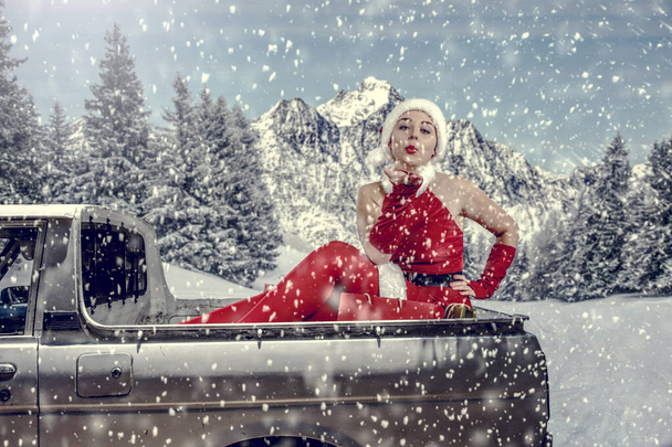 Woman Santa Claus in a car with snowy winter background. Xmas time and delivering presents. Santa Claus's female helper in a car driving to deliver some christmas presents on a sunny winter day. Blurred mountain forest background. - Photo, image