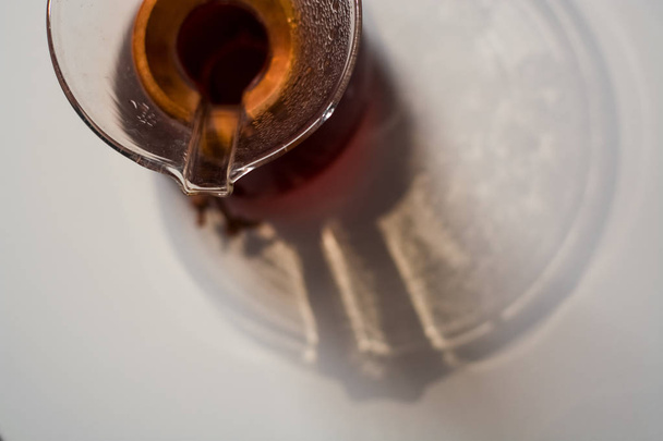 Γυάλινο δοχείο γεμάτο με μαύρο καφέ σε λευκό φόντο ενός τόνου με αντανάκλαση μεγάλων και ασυνήθιστων σκιών. - Φωτογραφία, εικόνα