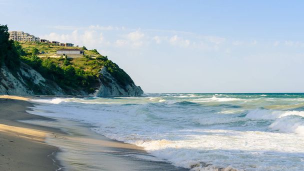 ondas do mar em uma praia arenosa contra um penhasco e céu azul
 - Foto, Imagem