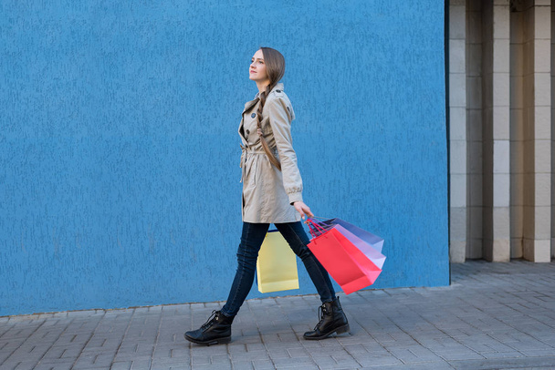 Happy young woman with colorful bags walking on the street. Blue wall on background - Fotó, kép