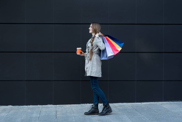 カラフルなバッグや紙コップを持つ幸せな若い女性。背景に黒い壁 - 写真・画像
