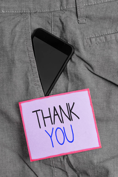 Σήμα που δείχνει "Ευχαριστώ". Εννοιολογική φωτογραφία μια ευγενική έκφραση που χρησιμοποιείται όταν αναγνωρίζετε μια συσκευή δώρου ή υπηρεσίας Smartphone μέσα σε επίσημο παντελόνι εργασίας μπροστινή τσέπη κοντά σε χαρτί σημειώσεων. - Φωτογραφία, εικόνα