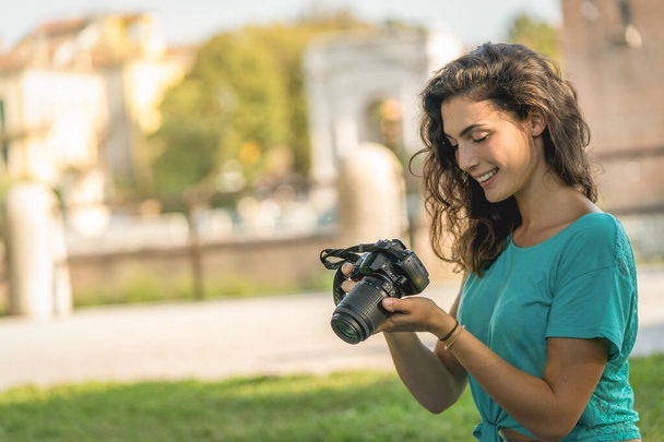 写真家の女の子は芝生の上でカメラの画面を見て、笑って座っている - 写真・画像