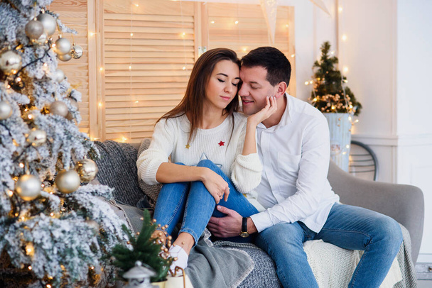 Attractive cheerful man and woman sitting near christmas tree and hugging each other. New Years eve. - Photo, Image