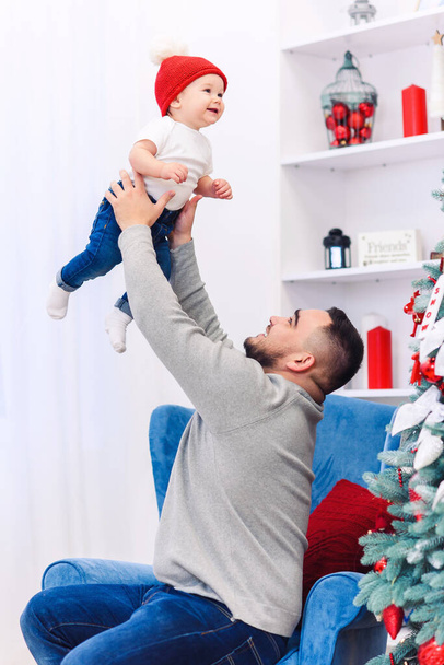 Handsome dad throwing up his amusing infant boy in the air near festive decorated christmas tree. - Foto, afbeelding