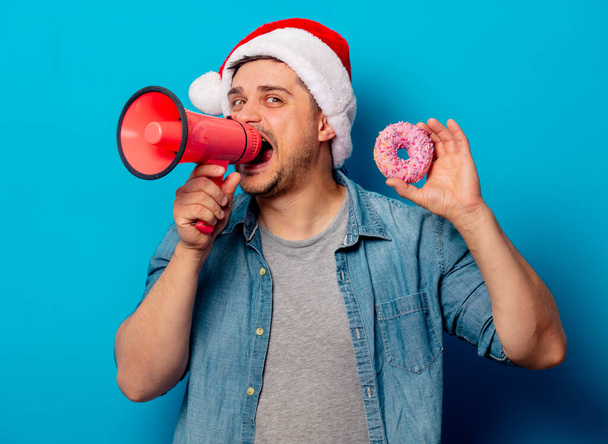 Knappe man met kerstmuts met donut en luide hailer op blauw - Foto, afbeelding