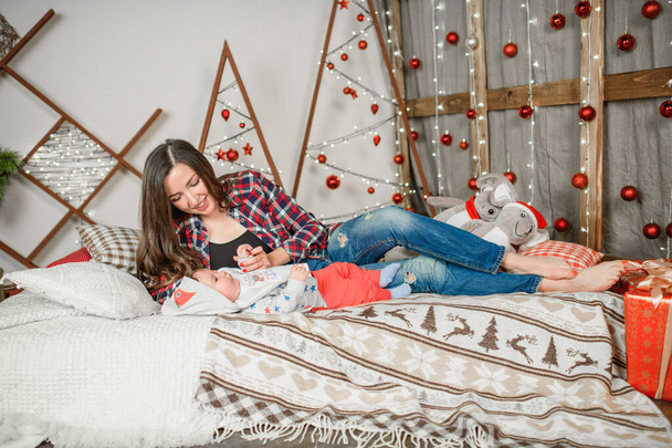 mladá matka s dítětem v novoroční atmosféře. Vánoční rodina, matka a dítě se usmívají u vánočního stromečku. Obývací pokoj je zdoben vánoční stromeček a dárková krabice, světlo gi - Fotografie, Obrázek