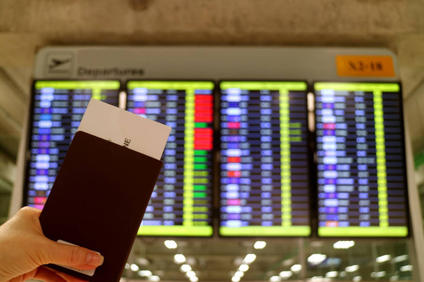 Паспорт с посадочным талоном в руке женщины с размытым экраном отображения информации о полете на заднем плане
 - Фото, изображение